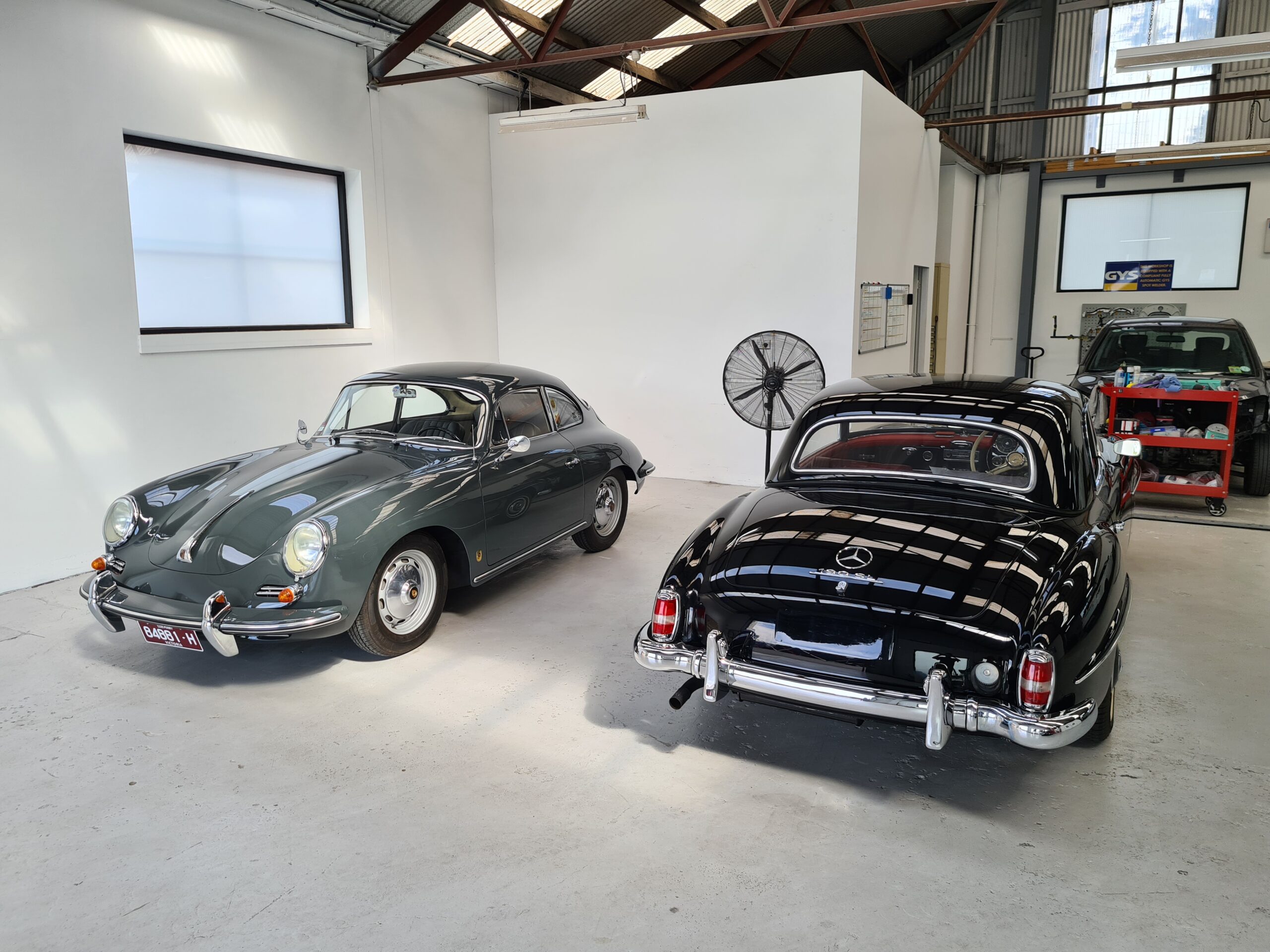 Two classic cars parked inside a well-lit garage with white walls and high ceilings. On the left is a dark green vintage Porsche 356 coupe, and on the right is a black Mercedes-Benz 190SL with a hardtop. A large industrial fan stands between the cars, and a modern black vehicle with a red tool cart is visible in the background.